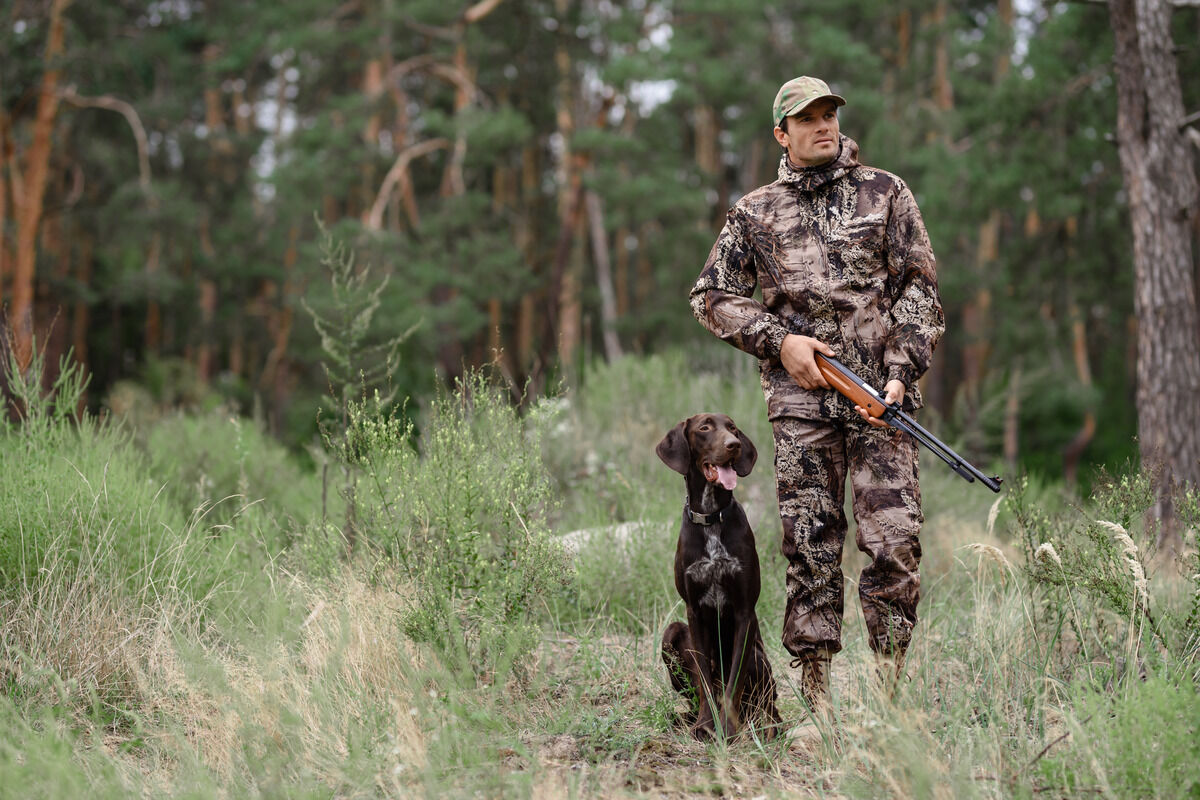 A hunter stands with his gun in the middle of the forest. He has full camouflage on and has a dog next to his leg.